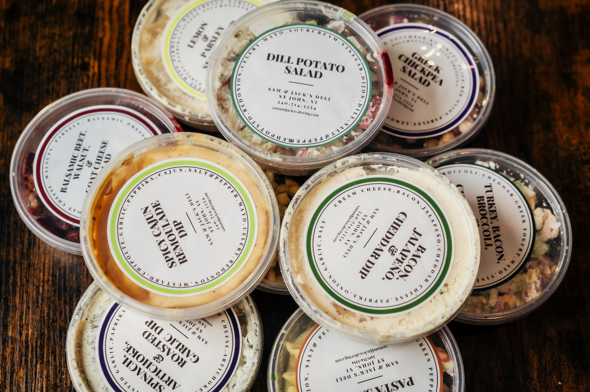 Stacked deli containers with labeled lids: dill potato salad, greek chickpea, spicy cajun remoulade, bacon jalapeño cheddar dip, balsamic beet, spinach & roasted garlic dip.